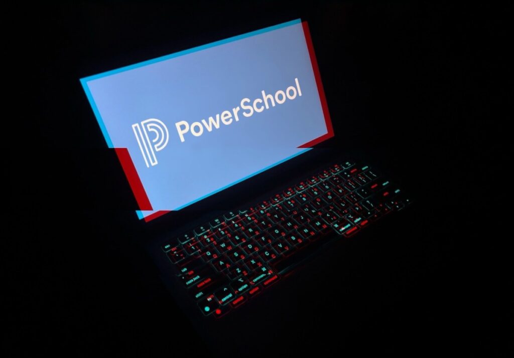 a laptop in a dark room with the laptop keyboard lit up with a red and blue glitchy effect, and the display reads PowerSchool with its corporate logo.