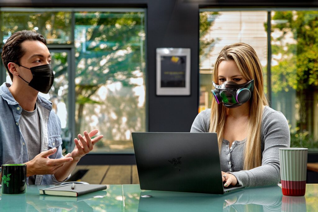 A person wearing a Razer Zephyr mask while typing on a laptop next to a person wearing a standard mask