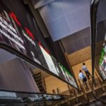 Pedestrians exit an escalator next to an electronic screen and ticker board displaying stock figures in Singapore