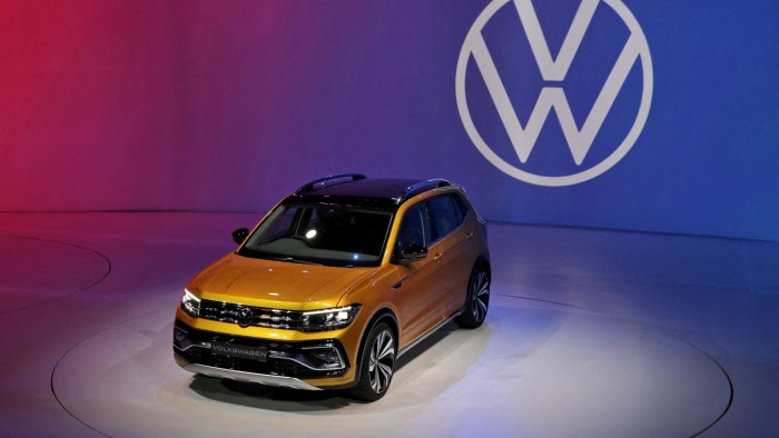 A yellow Volkswagen Taigun compact sport utility vehicle on display at an event with a gradient background transitioning from red to blue and the Volkswagen logo