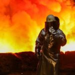 A worker in protective gear stands beside a blast furnace, with bright orange and yellow flames illuminating the scene at a steel plant