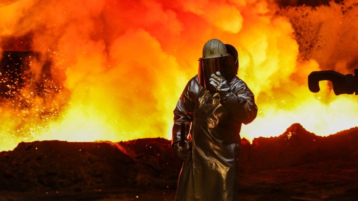 A worker in protective gear stands beside a blast furnace, with bright orange and yellow flames illuminating the scene at a steel plant