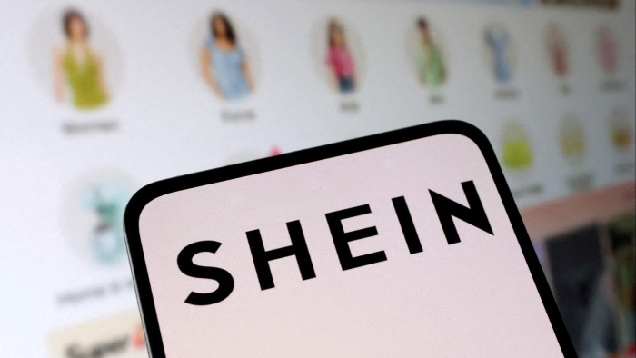 The Shein logo displayed on a smartphone is held in front of a screen displaying its web shop