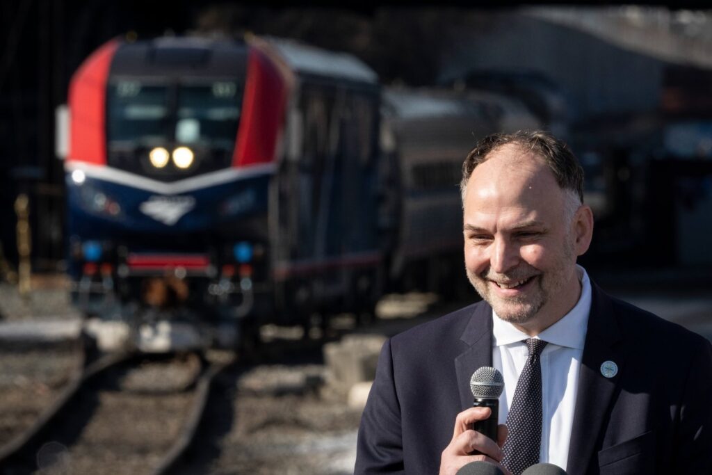 Amtrak CEO Stephen Gardner speaks into a microphone with a strip of track and an Amtrak train in the background