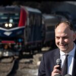 Amtrak CEO Stephen Gardner speaks into a microphone with a strip of track and an Amtrak train in the background