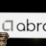 A toy figure of a businessman stands on a stack of pound coins, with the Abrdn logo displayed in the background.