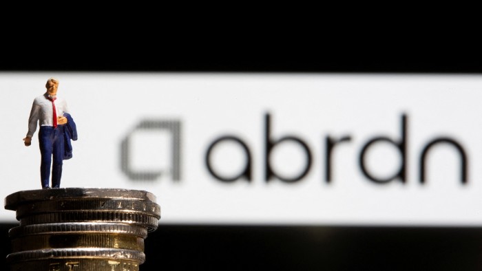 A toy figure of a businessman stands on a stack of pound coins, with the Abrdn logo displayed in the background.