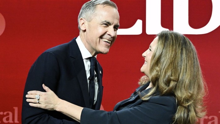 Mark Carney and Chrystia Freeland shake hands and smile warmly at each other against a red background