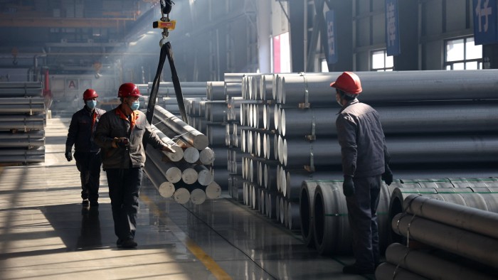 Workers in protective gear and red helmets operate a lift for aluminium rods in a factory filled with large stacks of rods and with sunlight filtering through the windows, illuminating the space