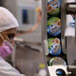 A worker in a hairnet and pink mask operates a production line at a Ben & Jerry's factory, with ice cream pots labelled &quot;Chunky Monkey&quot; visible on the conveyor