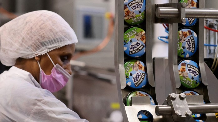 A worker in a hairnet and pink mask operates a production line at a Ben & Jerry's factory, with ice cream pots labelled &quot;Chunky Monkey&quot; visible on the conveyor