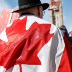 A man stands wrapped in a Canadian flag with his back facing the camera