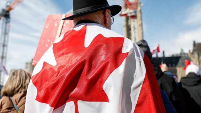 A man stands wrapped in a Canadian flag with his back facing the camera