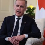 Mark Carney in a suit and sits in an armchair in front of a Canadian flag