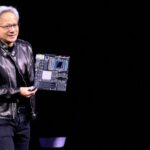 Jensen Huang holds up a circuit board while delivering a speech in a black leather jacket and standing against a dark background