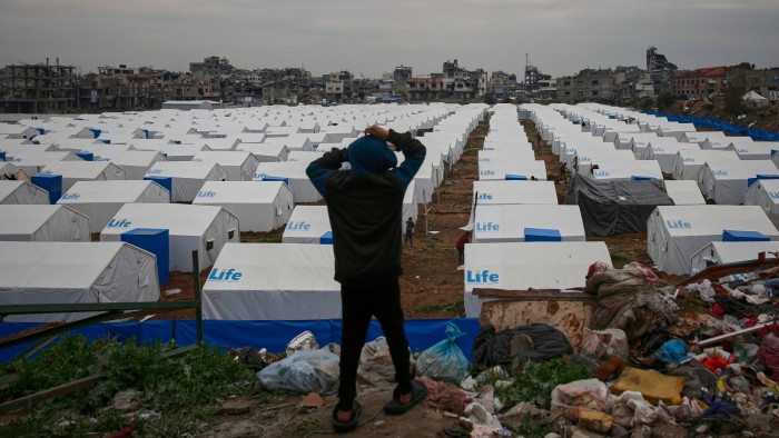 A boy stands on a hill, looking over a large tent camp for displaced Palestinians