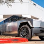 A Tesla Cybertruck is parked in front of a white dealership building in Austin, Texas. The vehicle is noted for its angular, futuristic design.