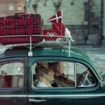 A couple in a green car with red luggage, a small pennant of the Danish flag and a red doll’s pram strapped to the roof, drive through a town